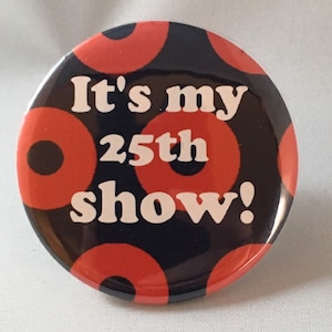 May include: A black and red button with white text that reads "It's my 25th show!" The button has a pattern of red circles with black centers.
