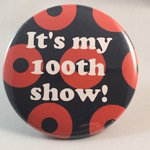 May include: A black and red button with white text that reads "It's my 100th show!" The button has a pattern of red circles with black centers.