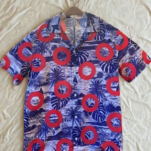 May include: Short-sleeved shirt with a tropical design. The shirt displays a pattern of red circles, palm trees, and large leaves against a blue and white backdrop. It is hanging on a wooden hanger.