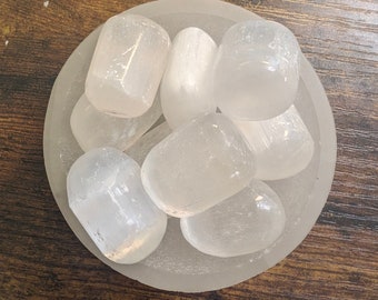 May include: A collection of smooth, white selenite tumbled stones in a clear glass bowl. The stones are various sizes and shapes, with a milky white color.