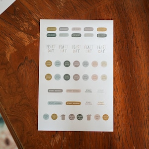 May include: A sheet of stickers with various text labels in different colours. Labels include "Birthday," "Feast Day," "Small Group," "Start Novena," and "Trash Day." The stickers are arranged in rows and columns on a white background.