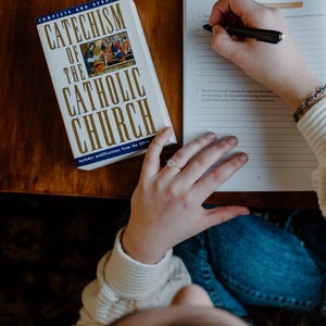 May include: A book titled "Catechism of the Catholic Church" with gold lettering and a colorful image. A person is writing on a notebook with a black pen. The scene is captured from an overhead perspective.