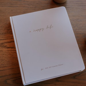 Peut inclure: Un agenda acad&eacute;mique blanc avec l'inscription dor&eacute;e "a happy life". L'agenda est pour l'ann&eacute;e scolaire 2026-2027. Un caf&eacute; glac&eacute; dans un gobelet transparent est pos&eacute; sur une table en bois en arri&egrave;re-plan.