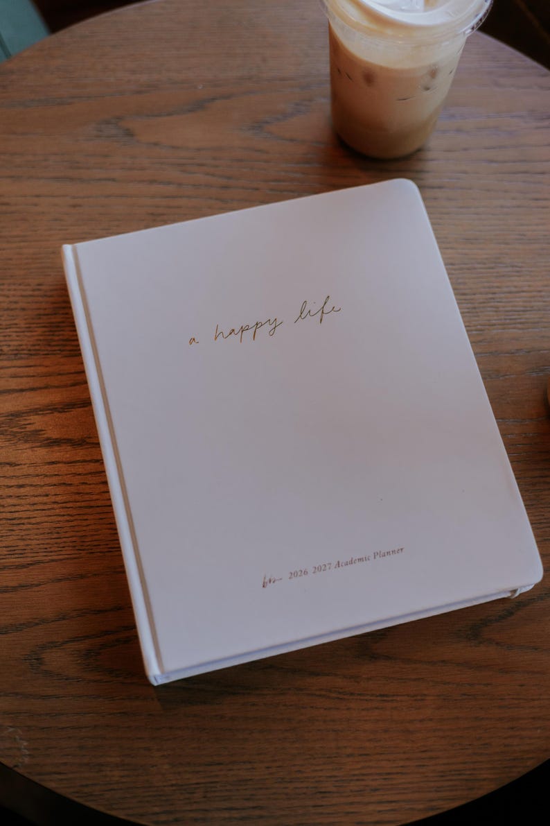 Peut inclure: Un agenda acad&eacute;mique blanc avec l'inscription dor&eacute;e "a happy life". L'agenda est pour l'ann&eacute;e scolaire 2026-2027. Un caf&eacute; glac&eacute; dans un gobelet transparent est pos&eacute; sur une table en bois en arri&egrave;re-plan.