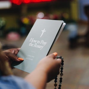 May include: A gray booklet titled "How to Pray the Rosary" with a cross symbol. The booklet is held with a rosary in the hand. The background is blurred, with hints of red and yellow.