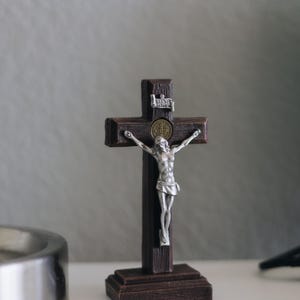 Puede incluir: Un crucifijo de madera marrón oscuro con una figura de Jesús plateada. La cruz tiene una base decorativa. La figura tiene los brazos extendidos. Un pequeño emblema metálico está en la parte superior de la cruz.