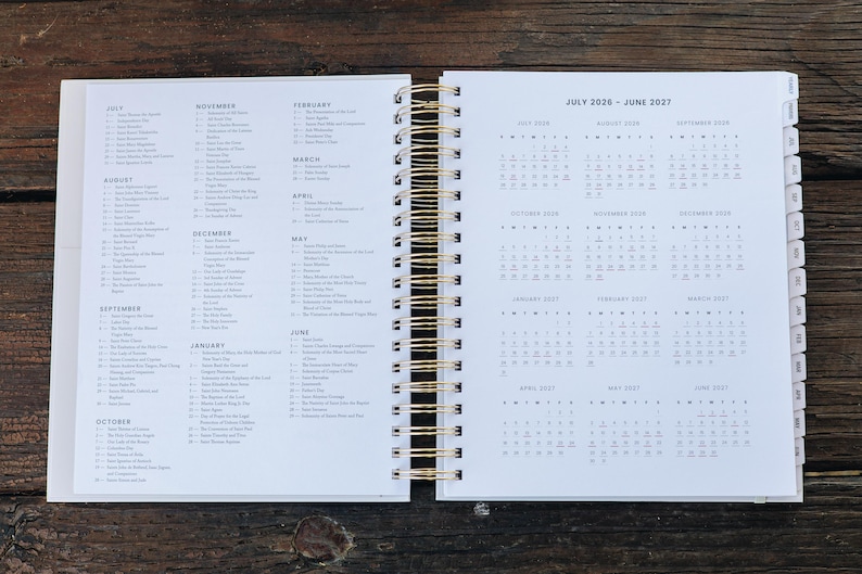 Peut inclure: Agenda ouvert avec des pages blanches et une reliure spirale dor&eacute;e. La page de gauche r&eacute;pertorie les mois avec les dates, tandis que la page de droite affiche un calendrier de juillet 2020 &agrave; juin 2021. L'agenda est ouvert sur une surface en bois fonc&eacute;.