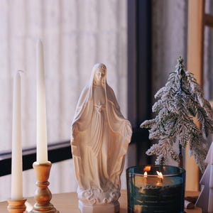 May include: A serene scene featuring a white statue of a religious figure, flanked by two white candles in wooden holders. A lit teal candle and a small frosted Christmas tree are also present on a wooden surface, creating a peaceful ambiance.