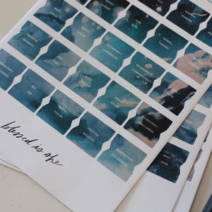 Op de afbeelding: Een stapel witte papieren vellen met blauwe tabbladen in aquarel-stijl. Elk tabblad heeft een titel. De zin "blessed is she" is in cursief geschreven in de linkerbenedenhoek. Rechts bevindt zich een donkerblauw object.