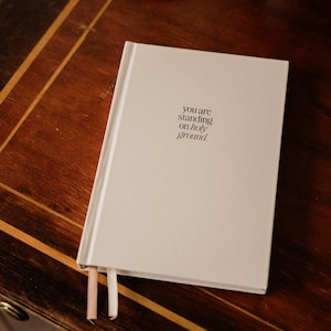 Könnte beinhalten: Ein weißes Journal mit dem Text "you are standing on holy ground." auf dem Einband. Das Journal liegt auf einer Holzoberfläche.