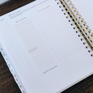 Peut inclure: Agenda ouvert avec une reliure &agrave; spirale dor&eacute;e, pr&eacute;sentant des pages lign&eacute;es et des sections pour la planification des repas et les listes de t&acirc;ches. La semaine du 7 juin est mise en &eacute;vidence, avec une citation en haut. Des onglets de mois sont visibles.