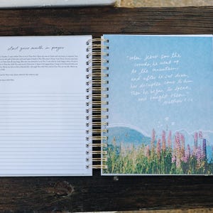 Peut inclure: Journal &agrave; spirales ouvert, avec des spirales dor&eacute;es. La page de gauche contient du papier lign&eacute; avec le texte "start your month in prayer". La page de droite a un fond bleu avec une illustration de paysage et du texte de Matthieu 5:1-2.