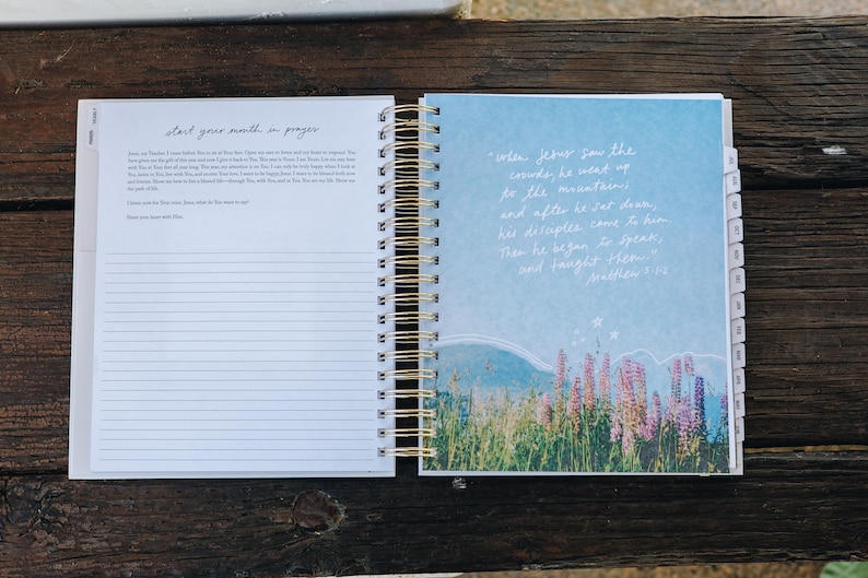 Peut inclure: Journal &agrave; spirales ouvert, avec des spirales dor&eacute;es. La page de gauche contient du papier lign&eacute; avec le texte "start your month in prayer". La page de droite a un fond bleu avec une illustration de paysage et du texte de Matthieu 5:1-2.