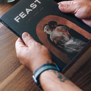 Puede incluir: Una persona sostiene un libro titulado "FEAST" con una cubierta oscura y letras blancas. El libro presenta una ilustración de dos figuras dentro de un arco. Un reloj de rayas azules y verdes es visible en la muñeca.