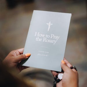 May include: A light gray booklet titled "How to Pray the Rosary" with a cross symbol. The booklet is held in hands with orange nail polish, accompanied by a rosary. The guide is from "blessed is she".