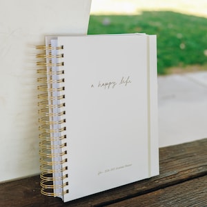 Peut inclure: Un agenda blanc &agrave; spirales dor&eacute;es. La couverture porte l'inscription "a happy life" en &eacute;criture cursive. L'agenda est pour l'ann&eacute;e scolaire 2026-2027. Il repose sur une surface en bois, avec un fond vert.