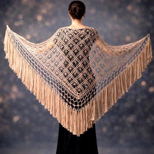 May include: A beige, open-weave shawl with a geometric pattern and long fringe. The shawl is draped over a person wearing a black dress. The background is a dark blue with bokeh lights.