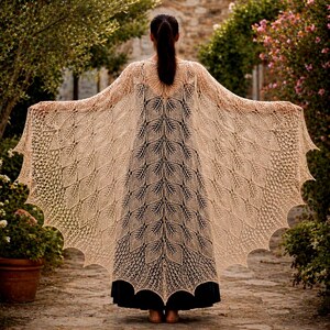 May include: A beige, open-weave shawl with a leaf-like pattern, draped over a black dress. The shawl has a scalloped edge and is spread wide, showcasing its intricate design. The background features a stone pathway and lush greenery.