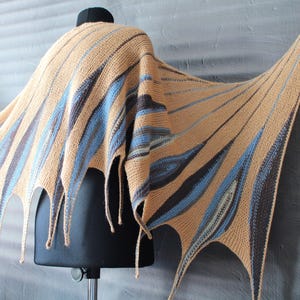 May include: A knitted shawl with a geometric pattern in shades of brown, blue, and beige. The shawl has a fringed edge and is draped over a mannequin.