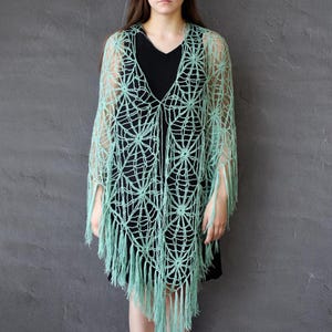 May include: A light green, open-weave shawl with a spiderweb design. The shawl has a V-neck and fringe along the bottom edge. The garment is worn over a black dress.
