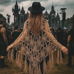 May include: A tan crocheted shawl with a spiderweb design and fringe. The shawl is worn over a black outfit, and a black hat is worn on the head. The background features a gothic-style building and a crowd of people.