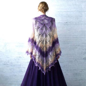 May include: A triangular lace shawl featuring an intricate leaf pattern in a gradient of purple, lavender, and cream colors. The shawl is draped over the shoulders, showcasing a delicate, open-knit texture with a pointed hem and scalloped edges.