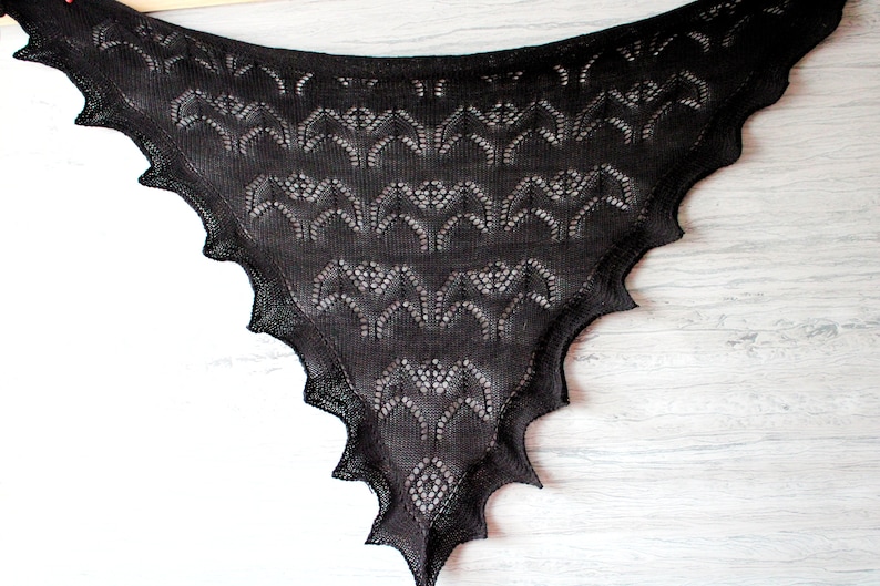 K&ouml;nnte beinhalten: Ein schwarzer gestrickter Schal mit einem Spitzenmuster aus Flederm&auml;usen. Der Schal ist dreieckig und hat einen gewellten Rand.