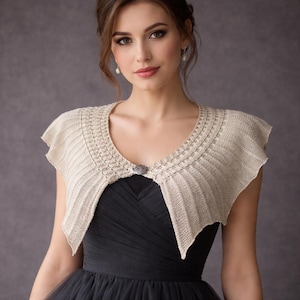 May include: A beige knitted shrug with a decorative neckline and a small, ornate clasp. The shrug is worn over a black dress, creating a layered look. The style is elegant and suitable for formal occasions.