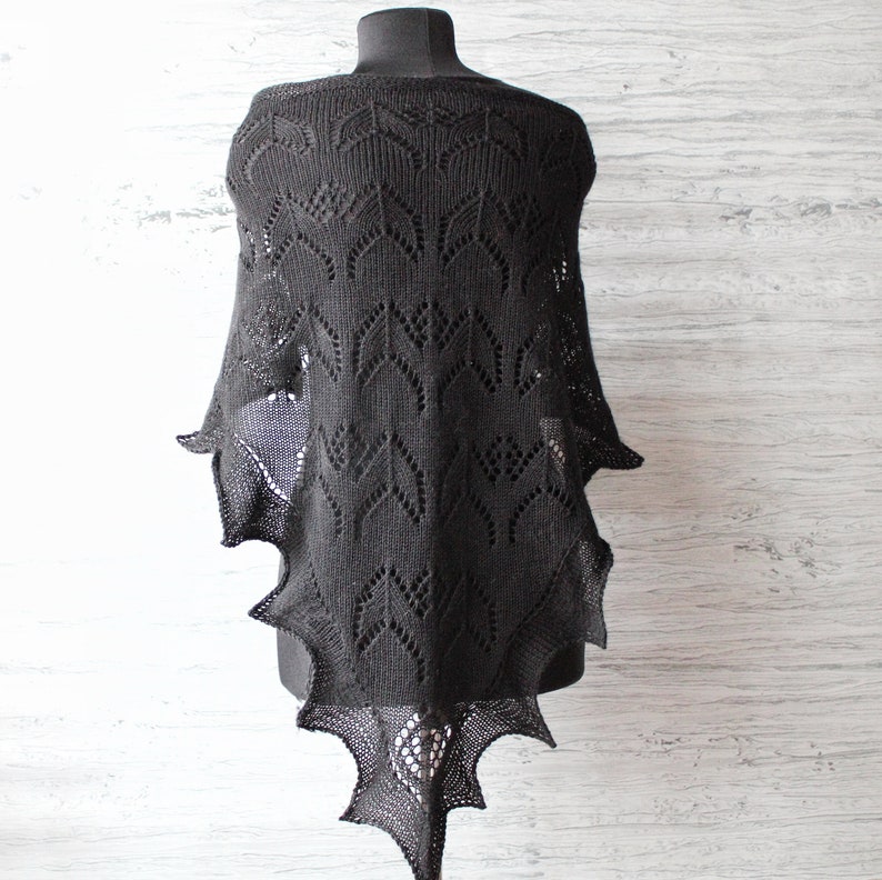 K&ouml;nnte beinhalten: Schwarzer gestrickter Schal mit einem zarten Spitzenmuster. Der Schal hat eine dreieckige Form mit einer spitzen Spitze und einem gewellten Rand.