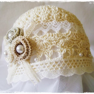 Puede incluir: Un sombrero de ganchillo con un delicado borde de encaje y adornos de perlas. El sombrero es de color crema y presenta un diseño floral.