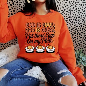 May include: Orange sweatshirt with the text "GOD IS GOOD, GOD IS GREAT, Put those Eggs On my Plate." The design includes three deviled eggs. The sweatshirt is a long-sleeved crew neck.