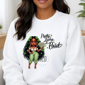 May include: White sweatshirt featuring a cartoon illustration of a woman with black curly hair adorned with green shamrocks. She holds money and wears green pants. The text "Pretty, Lucky, & Paid" is printed on the sweatshirt.