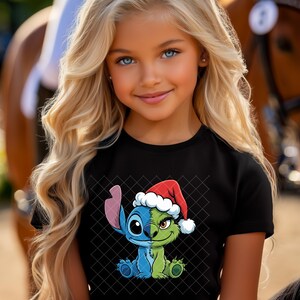 May include: A young girl with long blonde hair and blue eyes wearing a black t-shirt. The shirt features a split cartoon character design, half blue and half green, wearing a Santa hat. The background includes a horse.