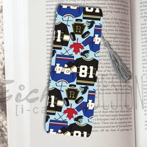 May include: A decorative bookmark featuring a pattern of hockey jerseys, hockey sticks, and pucks on a light blue background. The jerseys are black and blue with white numbers. A gray tassel is attached.