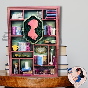 May include: A pink wooden miniature bookshelf with various decorative items. It features a silhouette, miniature books, a yellow cup, and glass jars. The shelf is on a wooden table, with books stacked in the background.