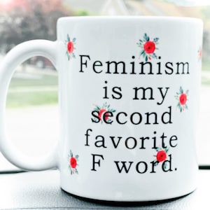 May include: White ceramic mug with a black inscription that reads "Feminism is my second favourite F word." The mug is decorated with small red flowers.