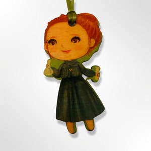 May include: A wooden ornament of a girl wearing a green dress and holding two small bottles. The girl has red hair and a smiling face.