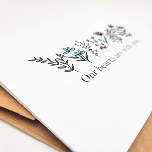 May include: A white card with a simple floral design and the text "Our hearts are with you".