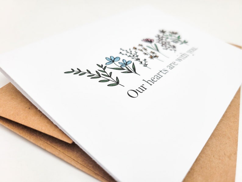 May include: A white card with a simple floral design and the text "Our hearts are with you".