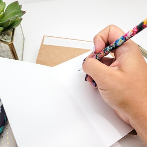 May include: A hand writing on a blank white card with a floral patterned pencil. The card is folded in half and is sitting on a table with a brown envelope and a small potted succulent.