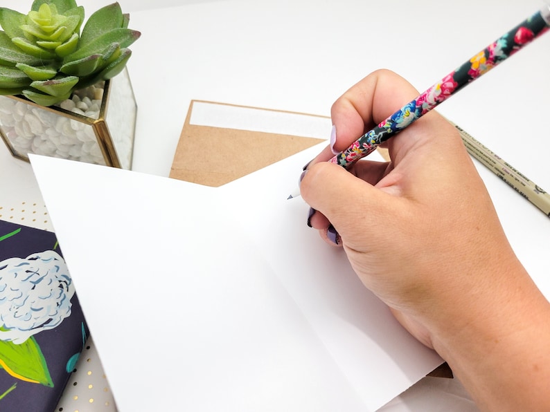 May include: A hand writing on a blank white card with a floral patterned pencil. The card is folded in half and is sitting on a table with a brown envelope and a small potted succulent.