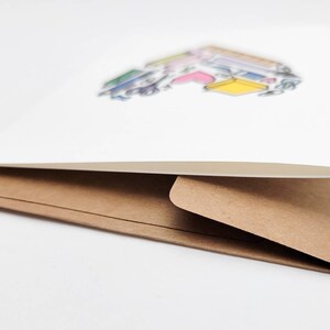 May include: A white card with a colorful illustration of books stacked on top of each other. The card is folded in half and has a brown envelope.