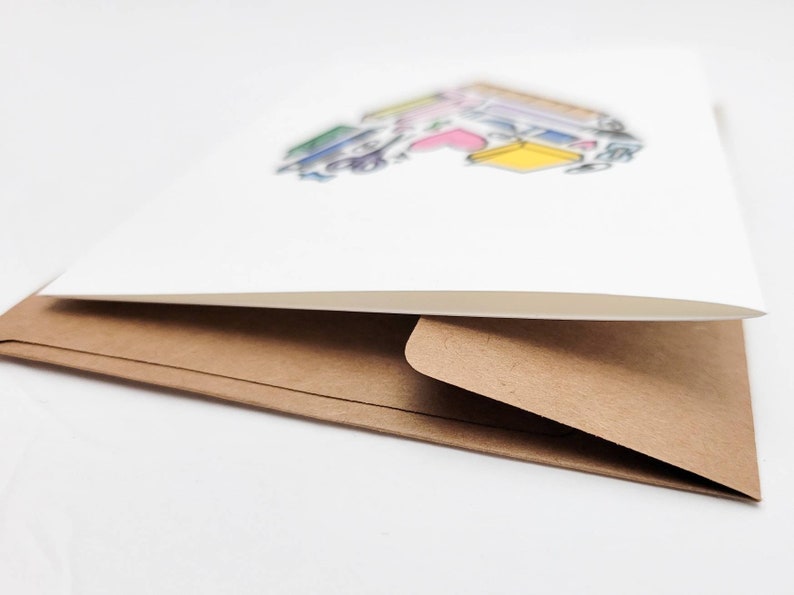 May include: A white card with a colorful illustration of books stacked on top of each other. The card is folded in half and has a brown envelope.