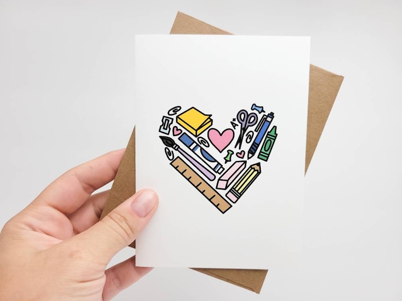 May include: A white card with a brown envelope. The card has a heart shape made of various school supplies, including a pencil, ruler, scissors, eraser, and a pink heart.