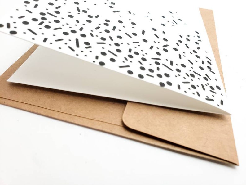 May include: A white card with a black and white geometric pattern is folded in half and placed on a brown envelope.