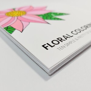 May include: A stack of colouring books with a pink flower and green leaves on the cover. The title of the book is "Floral Colouring: Ten Simple & Pretty Designs".