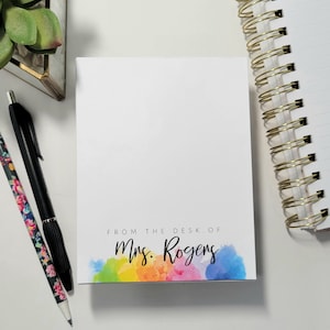 May include: White notepad with the text "FROM THE DESK OF Mrs. Rogers" in black script. The bottom edge features a watercolor design in rainbow colors. A black pen, a floral pencil, and a spiral notebook are also visible.