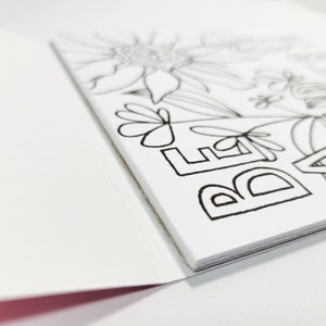 May include: A white card with black ink outlining the word "Believe" and floral designs. The card is folded in half.