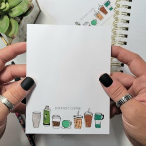 May include: A white notepad with the text "But First, Coffee" printed on it. The notepad features a row of illustrated coffee cups, including a green can, a glass, a paper cup, a mug, a cup with a lid, and a glass with a straw.