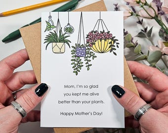 May include: A white card with a humorous Mother's Day message. The card features a cartoon illustration of three hanging plants with different colored pots. The text reads: "Mom, I'm so glad you kept me alive better than your plants. Happy Mother's Day!"